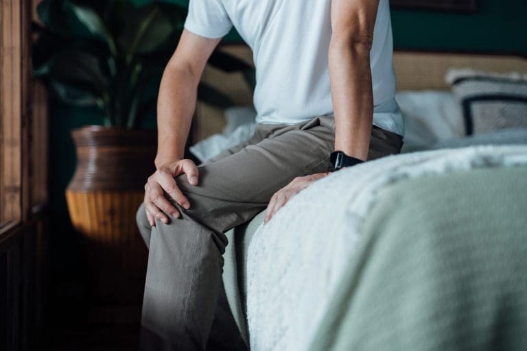 Close Up Of Senior Man Holding His Knee In Discomfort Suffering From Knee Pain While Sitting On Bed At Home Elderly And Health Issues Concept